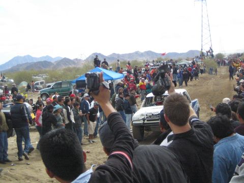 1990 Ford Bronco - white off-road race truck passes a dense crowd raising camcorders along a dirt desert course backed by mountains.
