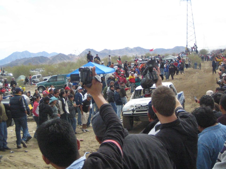 White off-road truck driving past crowds with raised cameras on a desert course