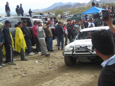 1990 Ford Bronco - white off-road truck fitted with a brush guard and roof lights sits on sandy ground near spectators and parked vehicles.