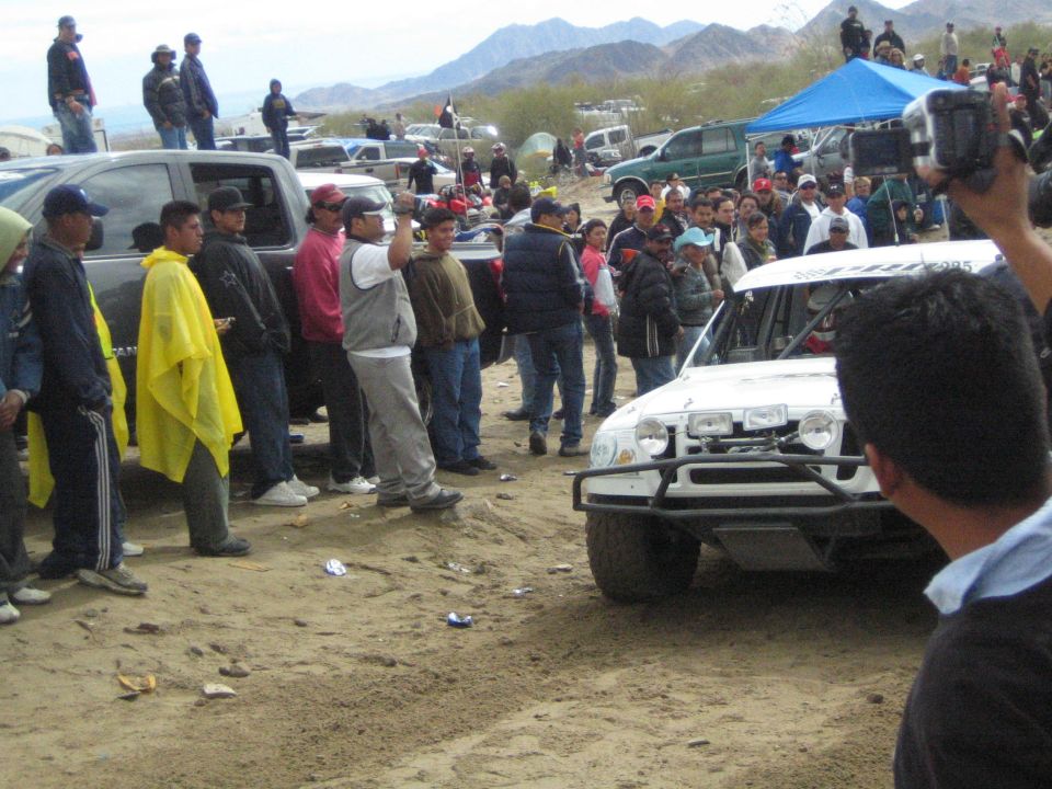 White off-road truck with brush guard parked near spectators on desert sand
