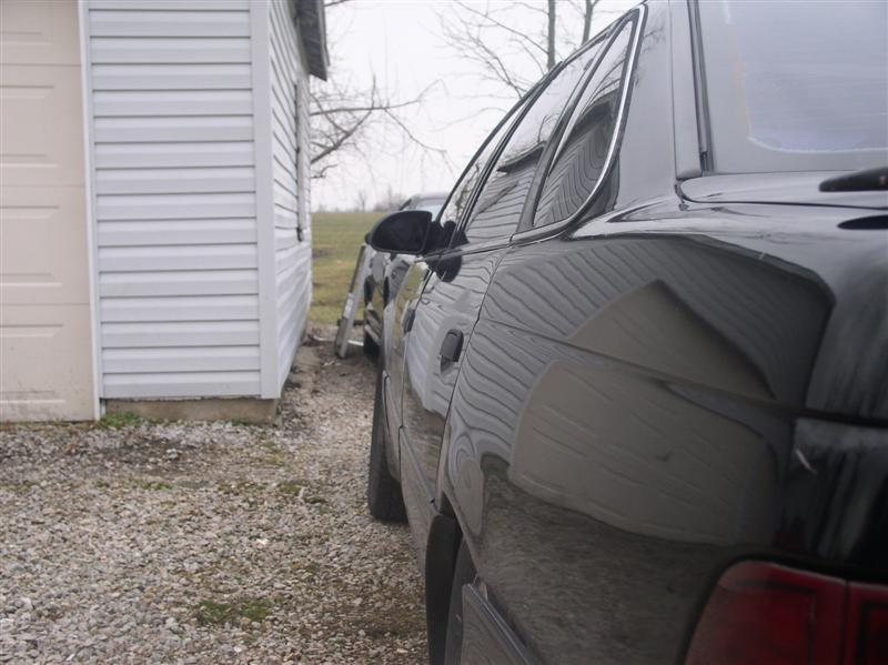 Black sedan parked next to a white building, showing its side profile and rear quarter panel