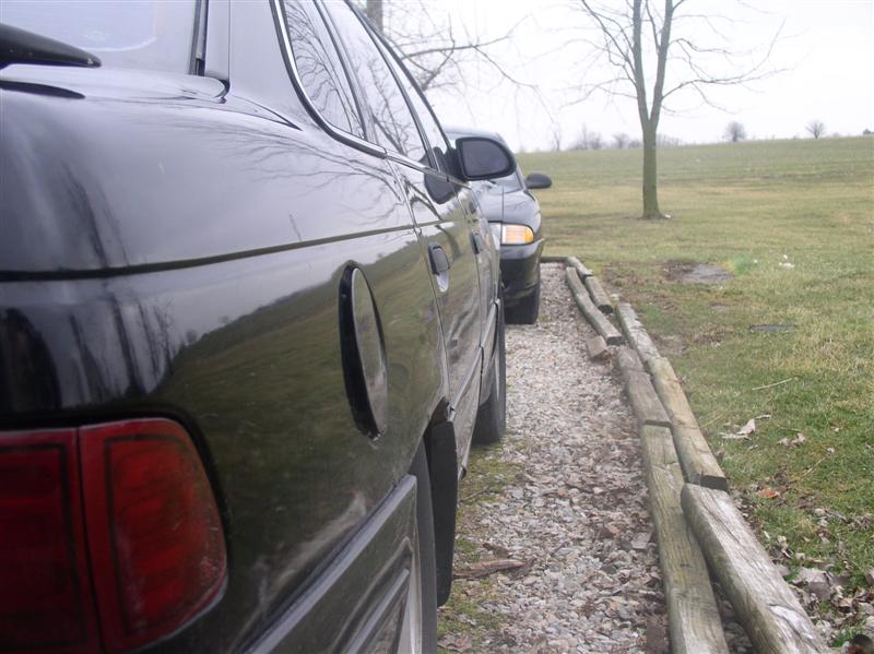 Side view of a black 1997 Ford Taurus and another car parked on gravel