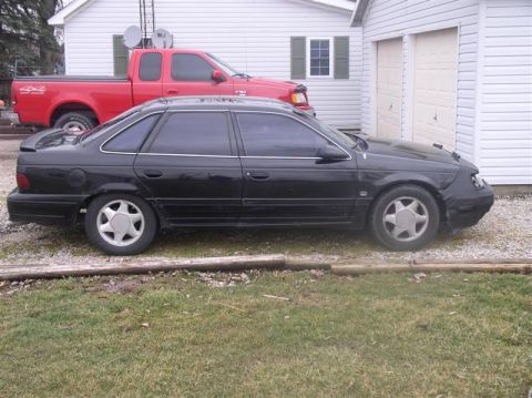1997 Ford Taurus - A black sedan is parked in front of a garage, with a red pickup truck behind it.