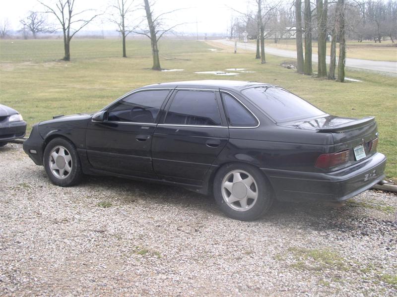 Black sedan parked on gravel, side view. Another car is partially visible to the left