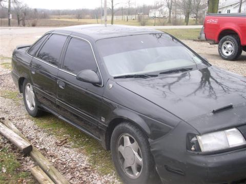 1997 Ford Taurus - A black sedan is parked on a gravel lot with a red pickup truck visible in the distance.