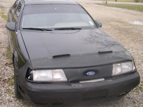 1997 Ford Taurus - A black Ford Taurus with a car cover is parked outdoors, showing a damaged headlight and tinted windshield.