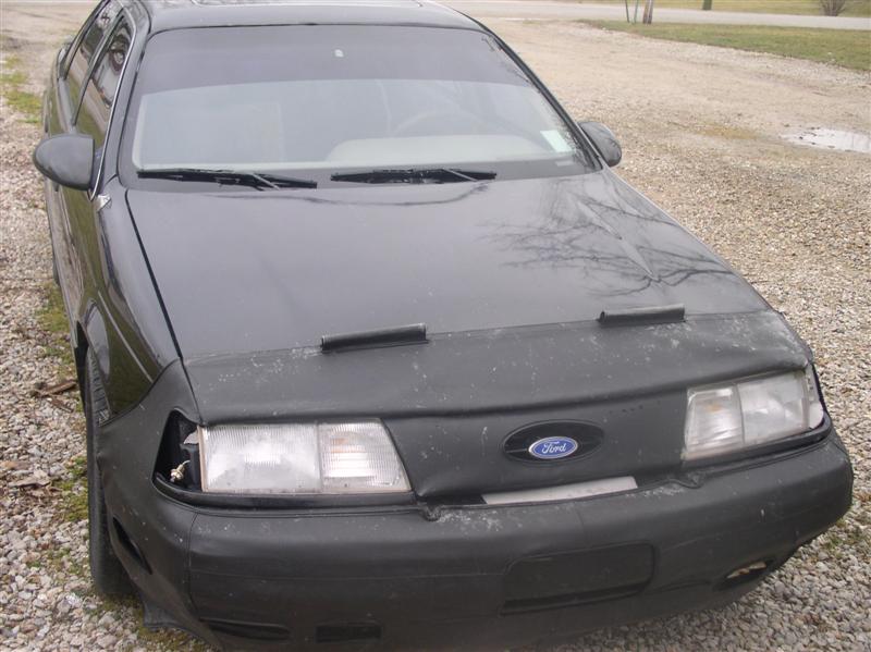 Black Ford Taurus with a car cover, damaged headlight, and tinted windshield