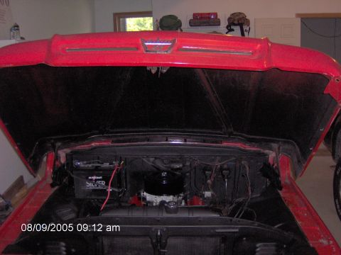 1960 Ford F-100 - The engine bay of a red pickup truck is visible with the hood raised, revealing the battery and engine components.