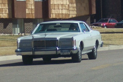 1972 Ford Ranchero - A white 1977 Mercury Grand Marquis sedan is seen from the front, driving on a street with other vehicles in the background.