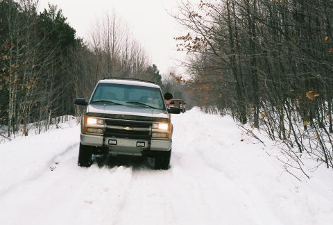 2000 Chevrolet Tahoe - Two SUVs drive down a snowy path lined with bare trees.
