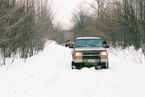 2000 Chevrolet Tahoe - Multiple trucks navigate a snow-covered path surrounded by bare trees.