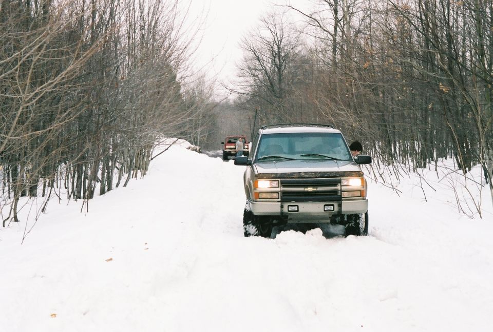 Several trucks driving down a snow-covered road through trees. The lead truck is a light-colored pickup