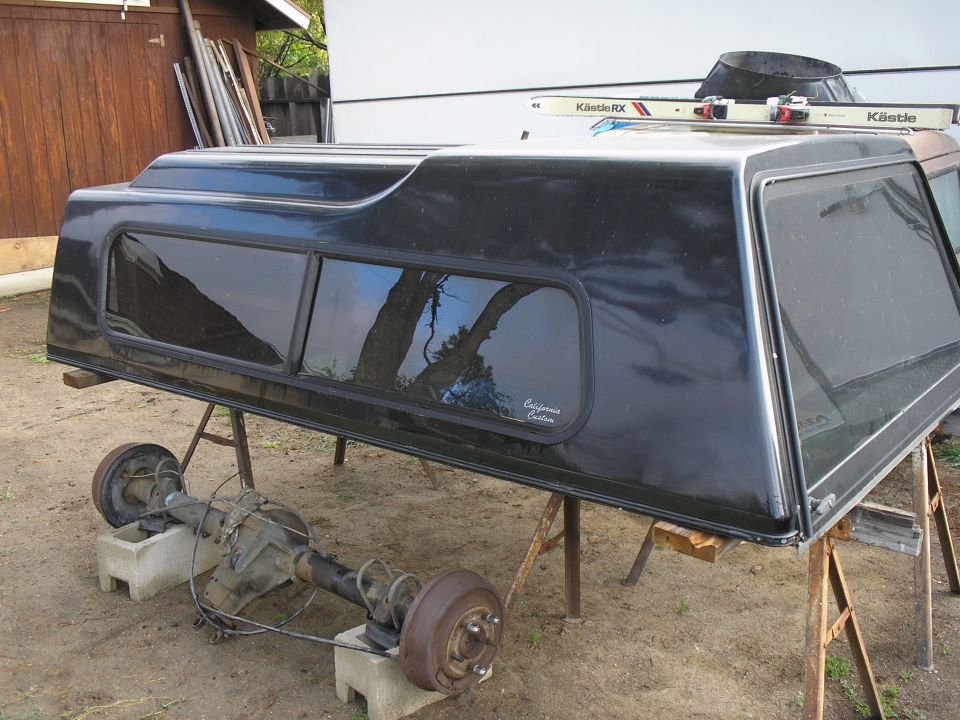 Black truck cab-high canopy shell on sawhorses next to a bare axle in a dirt yard