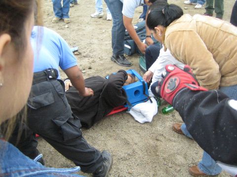 1990 Ford Bronco - person lies on the sand wearing a blue head-stabilization block while medics and onlookers attend to them at an outdoor event.