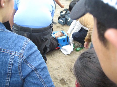 1990 Ford Bronco - overhead view of a patient immobilized with a blue head block on the sand, encircled by standing spectators and a paramedic.
