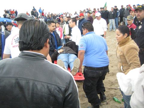 1990 Ford Bronco - Rescue workers in blue carry a person strapped to a red backboard stretcher through a dense crowd at a desert event.