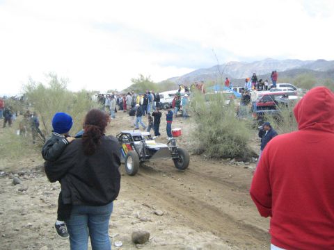 1990 Ford Bronco - open-frame off-road sand rail drives down a dirt trail lined with spectators against a backdrop of desert mountains.