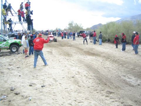 1990 Ford Bronco - red off-road buggy speeds along a sandy desert course as spectators line the route and climb a scaffold tower.