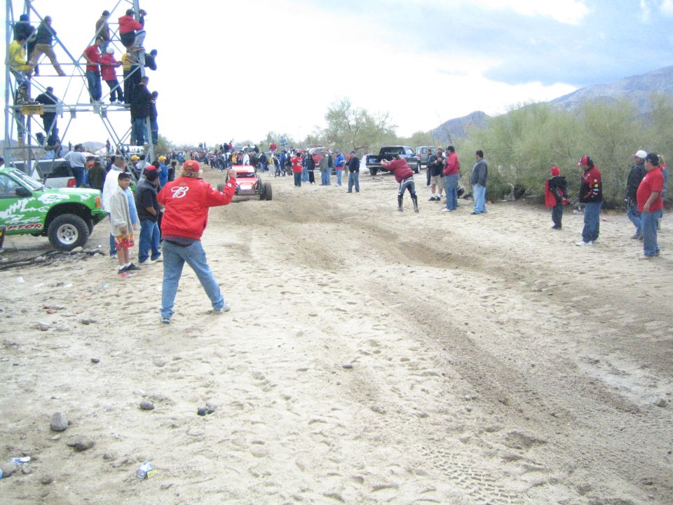 Red off-road buggy on sandy desert course with crowds and a green truck at a tower