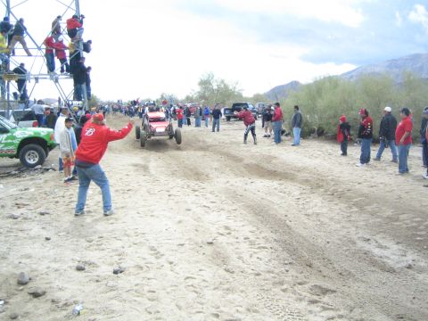 1990 Ford Bronco - red off-road racing buggy churns through deep sand as crowds watch from the trailside and a metal scaffold tower.
