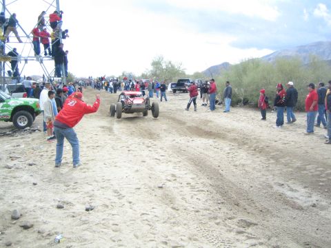 1990 Ford Bronco - red open-wheel off-road buggy maneuvers across the sandy course while a green truck and many spectators line the trail.
