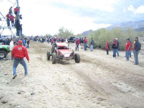 1990 Ford Bronco - red-and-white off-road buggy drives toward the camera on a sandy desert trail lined with spectators and distant mountains.
