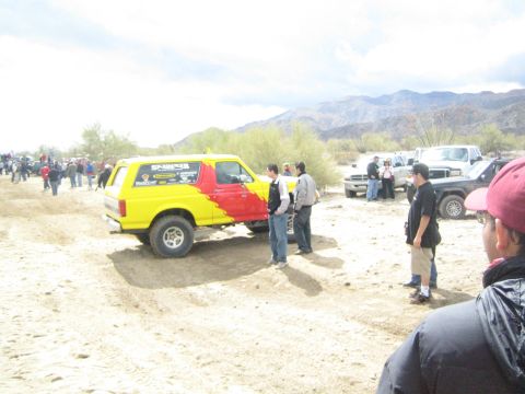 1990 Ford Bronco - yellow-and-red lifted SUV on mud tires sits on the sand surrounded by spectators at a desert off-road gathering.