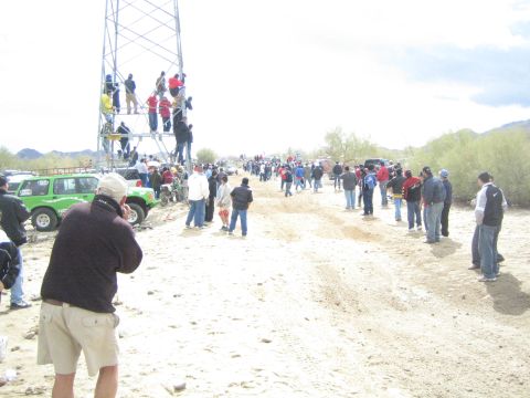1990 Ford Bronco - Crowds of spectators line a wide sandy desert trail, some climbing a metal scaffold tower, beside a green off-road truck.