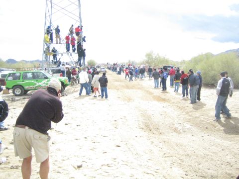 1990 Ford Bronco - man in cargo shorts photographs the action along a sandy desert trail crowded with spectators near a tall scaffold tower.