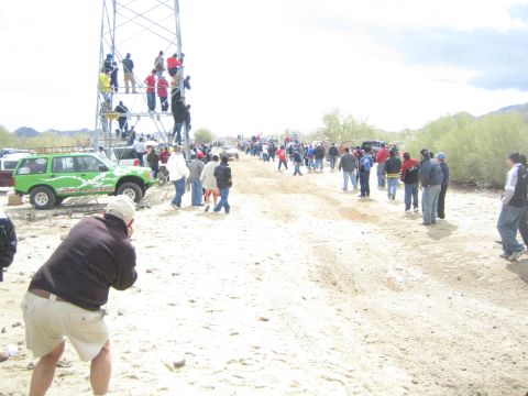 1990 Ford Bronco - green off-road truck sits trailside as spectators line a sandy desert course and others climb a scaffold tower.