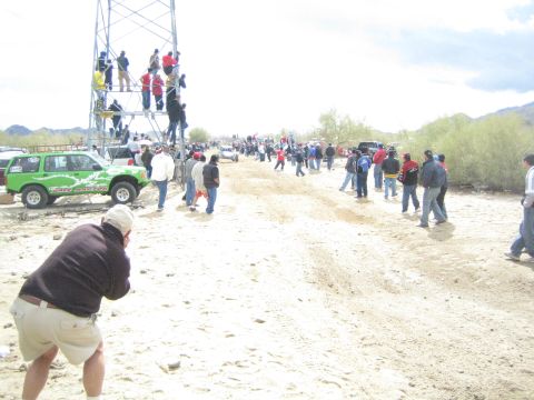 1990 Ford Bronco - bright green off-road SUV is parked beside a crowded sandy trail while spectators gather and climb a metal tower.