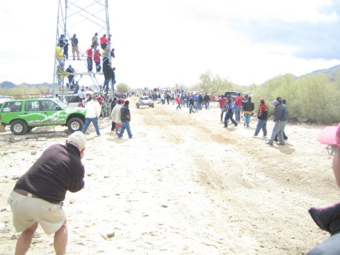 1990 Ford Bronco - Spectators in a desert line the sandy trail watching a distant off-road vehicle, with a green SUV and scaffold tower at left.