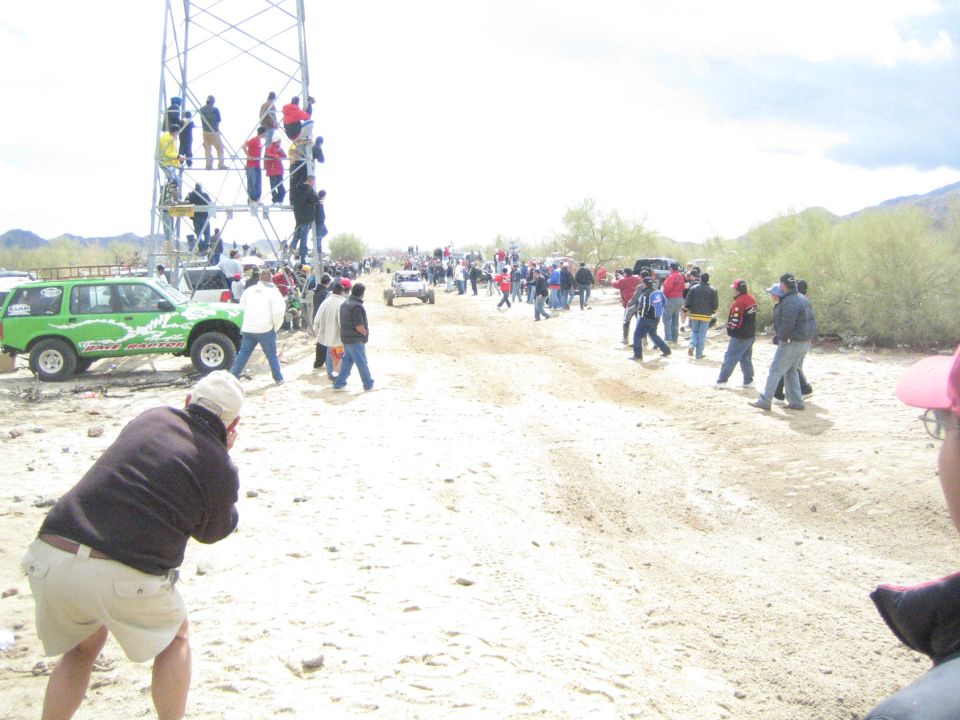 Spectators watch a distant off-road vehicle on a sandy trail near a scaffold tower