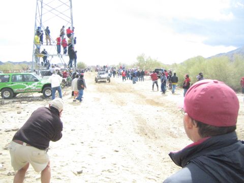 1990 Ford Bronco - small off-road vehicle approaches in the distance on a sandy desert trail as spectators and a man in a red cap watch.
