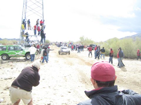 1990 Ford Bronco - off-road buggy heads down a sandy desert trail lined with spectators, a green SUV at left and people atop a scaffold tower.