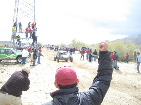 1990 Ford Bronco - spectator in a red cap raises a fist while an off-road buggy travels the sandy desert course past crowds and a scaffold tower.