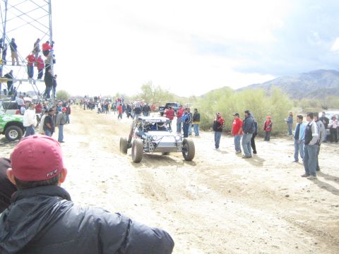 1990 Ford Bronco - silver tube-frame off-road buggy drives down the sandy desert trail as spectators line both sides under a cloudy sky.