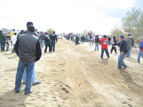 1990 Ford Bronco - large crowd of spectators stands along a wide sandy desert trail watching distant activity under an overcast sky.