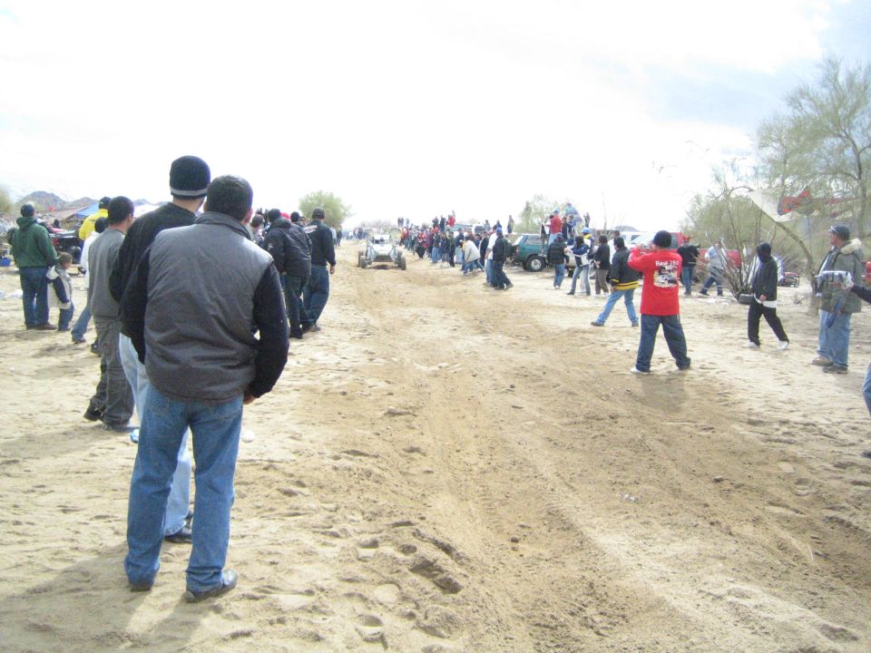 Spectators in winter jackets watch a distant off-road vehicle on a sandy trail