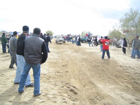 1990 Ford Bronco - crowd in winter clothing lines the sandy desert trail as a distant off-road vehicle and parked trucks sit down the course.