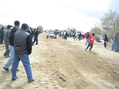 1990 Ford Bronco - Bundled spectators line the sandy desert trail watching a distant off-road buggy and parked trucks under a hazy sky.