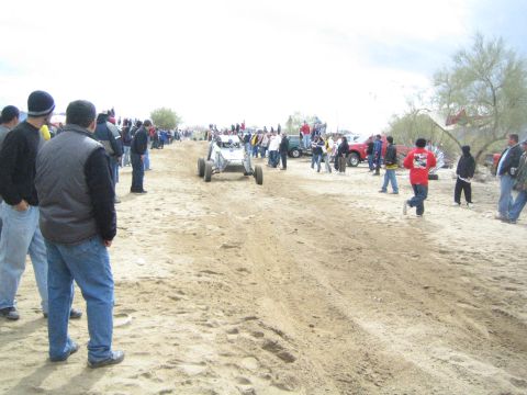1990 Ford Bronco - silver tube-frame off-road buggy sits on the sandy desert trail surrounded by spectators and parked trucks.