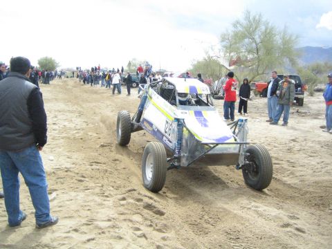 1990 Ford Bronco - white-and-blue tube-frame off-road buggy climbs a sandy berm on the desert course as spectators watch from the sidelines.
