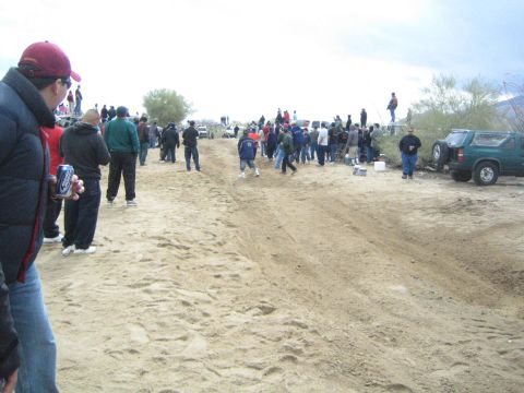 1990 Ford Bronco - Spectators gather along a sandy desert trail near a green SUV, watching distant activity beneath an overcast sky.