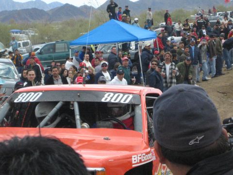 1990 Ford Bronco - red off-road race truck marked 800 with a BFGoodrich windshield band idles before a large crowd of spectators under a blue canopy.