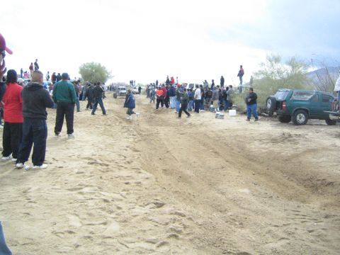 1990 Ford Bronco - Spectators gather along a churned sandy desert race course, with a green SUV and other vehicles parked at the trackside.