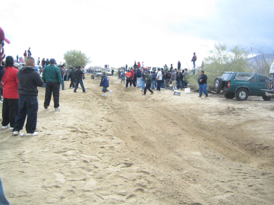 Spectators lining a sandy desert race course with parked SUVs and trucks