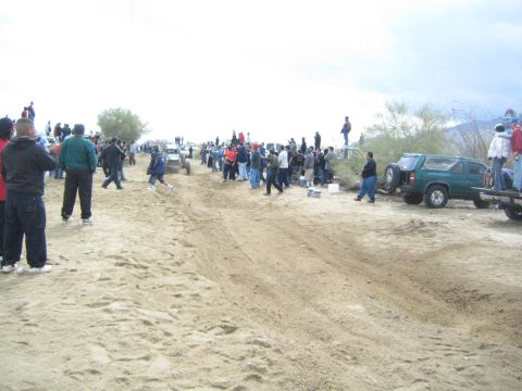 1990 Ford Bronco - wide sandy desert track lined with spectators, with a green Jeep Cherokee and other trucks parked along the edge.