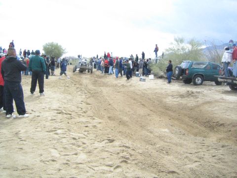 1990 Ford Bronco - open-cockpit off-road sand buggy works down a sandy desert track lined with spectators and parked trucks.