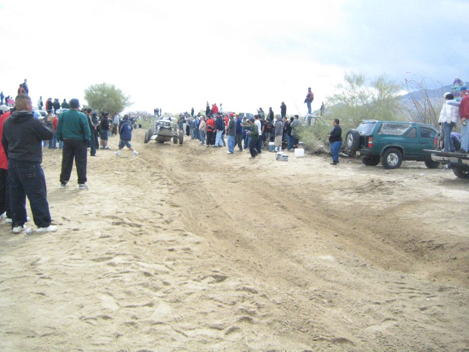 Open-cockpit off-road buggy on sandy track with spectators and parked green SUV